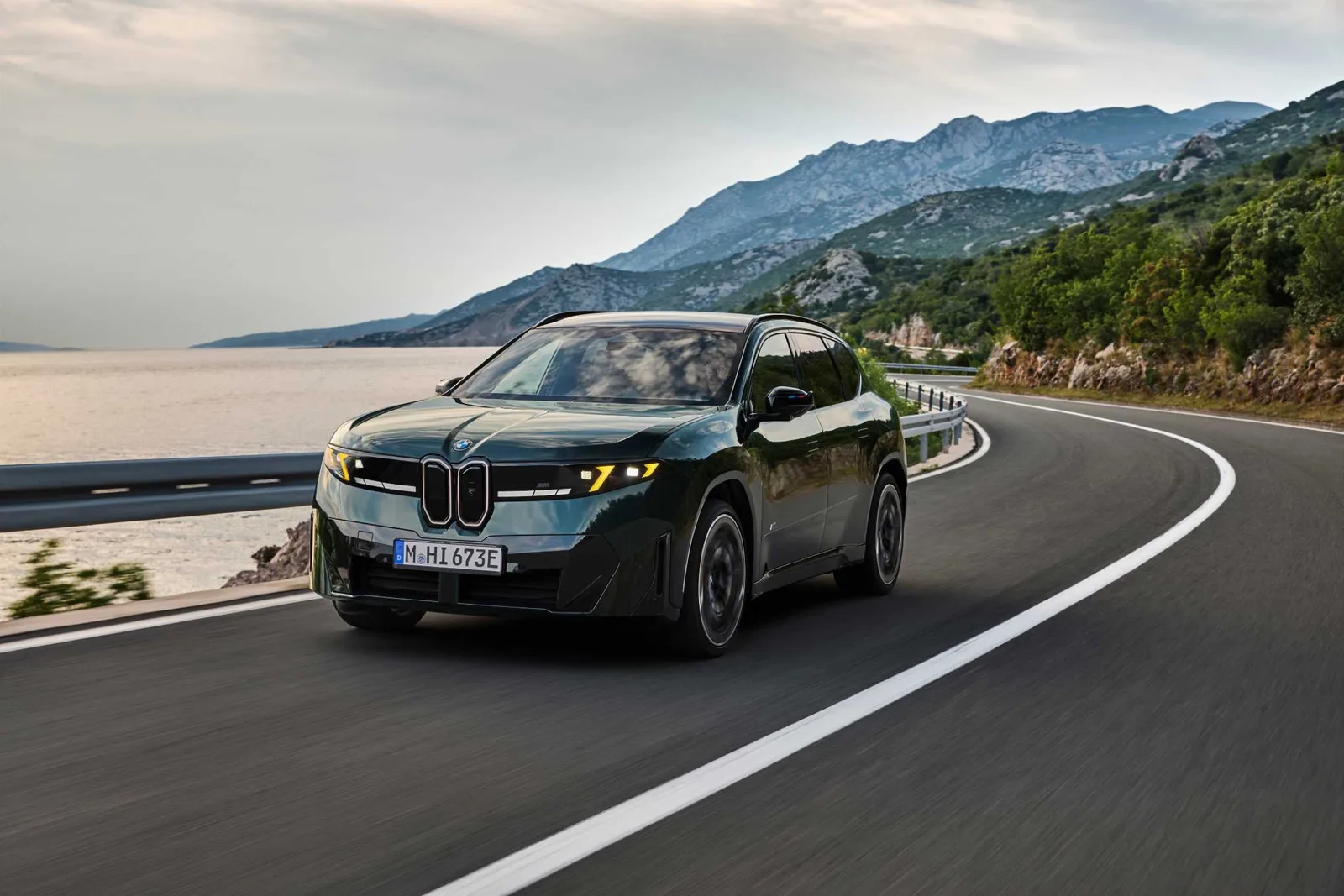 Donkergroene BMW iX3 M60 xDrive rijdend over een kustweg met bergen op de achtergrond