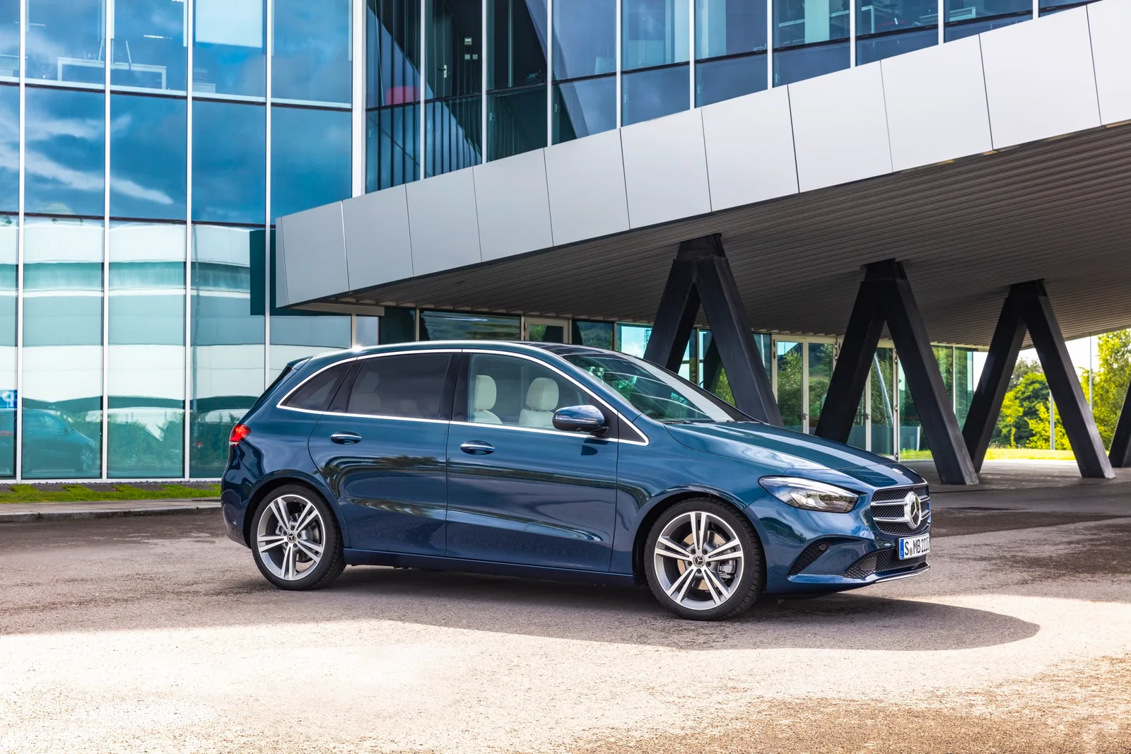 Donkerblauwe Mercedes-Benz B-Klasse van de W247-generatie voor een glazen gebouw