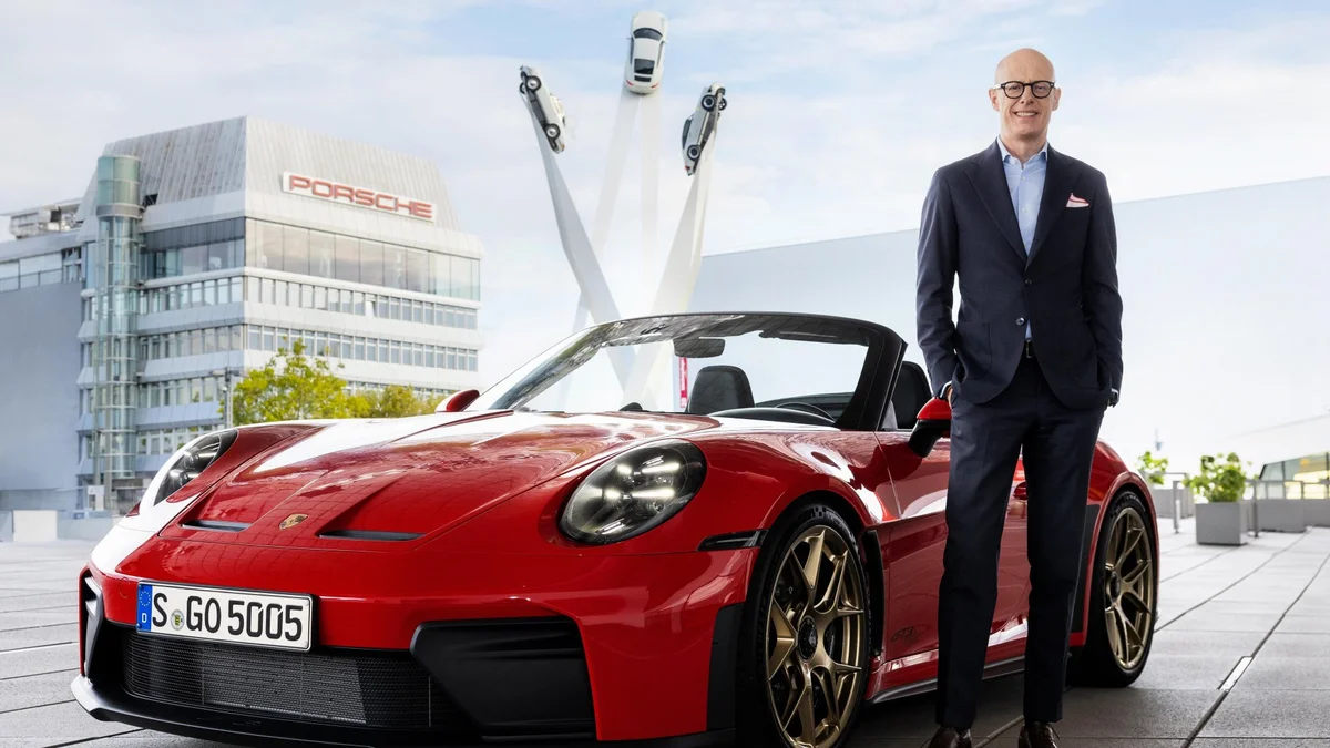 Rode Porsche 911 GT3 S/C met open dak en een man in pak, voor het Porsche-gebouw en het kunstwerk met drie Porsche 911's in Zuffenhausen